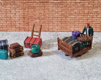 May include: A miniature wooden luggage cart with a person figure pushing it. The cart is loaded with various miniature suitcases in shades of brown, purple, and blue. There are also miniature suitcases and a ladder on the floor.