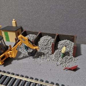 Puede incluir: Una escena de construcción en miniatura con una retroexcavadora amarilla y negra cargando grava en contenedores de madera. Hay una pequeña figura con una camisa amarilla. También se ven vías de tren en miniatura y pequeños edificios.