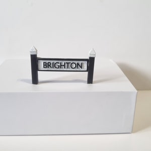 May include: A black and white sign with the word "BRIGHTON" printed on it. The sign is mounted on two black posts with white caps.