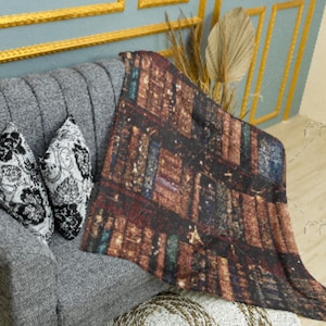 Puede incluir: Una manta con un diseño de libros de biblioteca sobre un sofá gris. Dos cojines decorativos con estampados florales en blanco y negro están en el sofá. La manta presenta una impresión detallada de estanterías con lomos de libros.