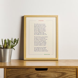 May include: A framed print of a poem in German, titled "Stufen" by Hermann Hesse. The poem is printed in blue and red ink on a cream-colored background. The frame is light wood and the print is sitting on a wooden shelf with two potted plants.