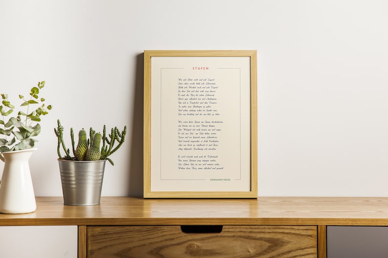 May include: A framed print of a poem in German, titled "Stufen" by Hermann Hesse. The poem is printed in blue and red ink on a cream-colored background. The frame is light wood and the print is sitting on a wooden shelf with two potted plants.