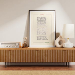 May include: A framed print of a poem titled "Stufen" by Hermann Hesse. The poem is printed in black ink on a white background. The frame is black and the print is hanging on a wall above a wooden console table. The console table has a white stereo receiver on the left side and a white lamp with a white shade on the right side. There is a small vase with dried flowers on the table.