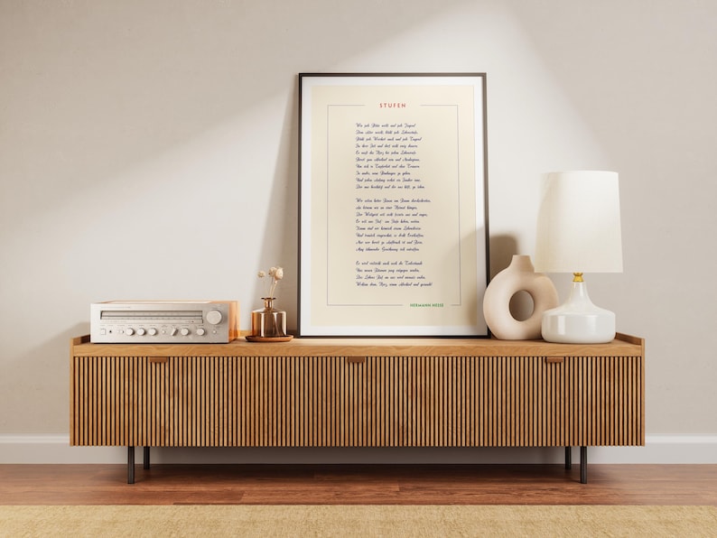 May include: A framed print of a poem titled "Stufen" by Hermann Hesse. The poem is printed in black ink on a white background. The frame is black and the print is hanging on a wall above a wooden console table. The console table has a white stereo receiver on the left side and a white lamp with a white shade on the right side. There is a small vase with dried flowers on the table.