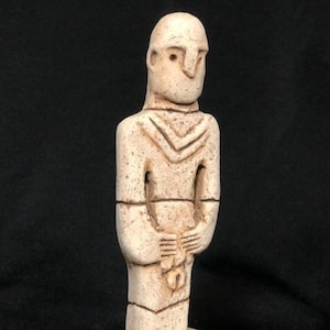 May include: A small, off-white statue of a standing figure with a stylized head and body. The figure has a geometric design on the chest and holds an object in its hands. It is mounted on a square base. The statue is approximately 15 cm tall.