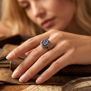 Könnte beinhalten: Ein goldener Ring mit einem kreisförmigen, tiefblauen Stein in einer Fassung. Der Stein ist mit vertikalen Linien und einem horizontalen Balken an der Oberseite graviert. Der Ring ist dünn und metallisch und hat ein poliertes Gold-Finish.