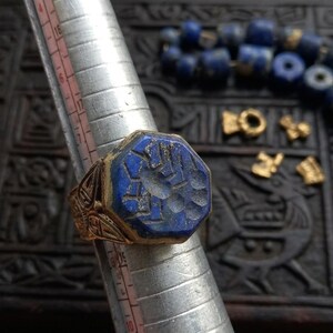May include: A gold-toned ring featuring a hexagonal lapis lazuli stone with an engraved design, positioned on a silver-colored metal ring mandrel marked with numerical sizing increments to display the band's diameter.