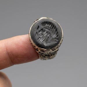 Könnte beinhalten: Ein silberner Ring mit einer kreisförmigen, tiefschwarzen Steineinlage, die ein eingraviertes, abstraktes geometrisches Muster aufweist. Das Metallband des Rings zeigt filigrane, detaillierte Schnitzereien, die den gesamten Umfang zieren.