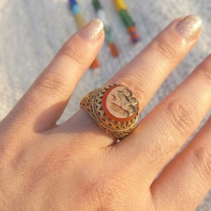 Könnte beinhalten: Ein Goldring mit einem komplexen Filigranband und einem zentralen, ovalen Karneolstein. Der Stein ist mit einem detaillierten Blumenmotiv graviert. Die Fassung hat einen gezackten Rand und das Metall zeigt ein feines, gewebtes Muster.