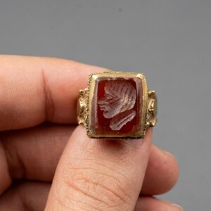 Könnte beinhalten: Ein Goldring mit einem quadratischen, tiefroten Karneol-Edelstein in der Mitte. Der Stein ist mit einem Profilporträt eines menschlichen Gesichts graviert. Der Goldring hat eine strukturierte, verzierte Oberfläche mit sichtbaren Metalldetails.