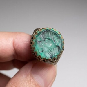 May include: A metal ring featuring a circular, translucent green stone inlay. The stone is engraved with a profile of a figure wearing a crested helmet. The metal setting has a scalloped edge and intricate, textured detailing along the band.