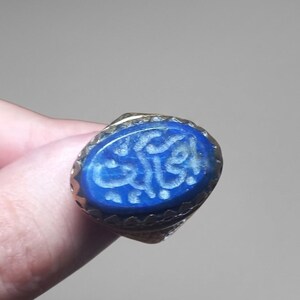 May include: A metal ring featuring an oval-shaped, deep blue lapis lazuli gemstone set in a serrated bezel. The surface of the stone is engraved with intricate, light-coloured Arabic calligraphy, creating a distinct contrast against the dark blue.