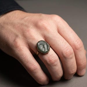 May include: A gold ring featuring an oval-shaped, dark green stone set in a bezel. The stone is engraved with a profile of a figure wearing a helmet, displaying intricate details on the surface of the gemstone. The band is thin and metallic.