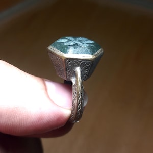 Könnte beinhalten: Ein Ring mit einem großen, sechseckigen, dunkelgrünen Stein in einer Metallfassung. Die Metallfassung und die Ringschiene sind mit komplizierten, eingravierten Mustern verziert. Der Stein hat eine texturierte, facettierte Oberfläche.