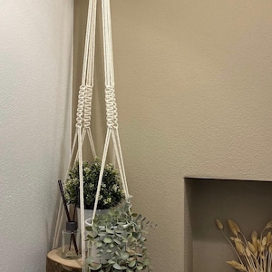 Pode incluir: Um suporte de plantas em macramé creme com uma prateleira de madeira que suporta uma planta em vaso e uma planta pendurada. Um pequeno difusor está na prateleira. O suporte está suspenso no teto.
