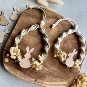 Op de afbeelding: Twee macramé kransen met houten konijn uitsparingen en gedroogde bloemen. Eén krans is lichtgroen, de andere is wit. Beide hebben een houten kraal aan de bovenkant. De kransen liggen op een houten dienblad.