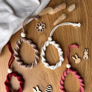 May include: Four macrame Easter egg decorations in red, beige, white, and pink, each with a wooden bead and hanging loop. Wooden bunny and egg cutouts are scattered on a wooden surface. The eggs are for Easter.