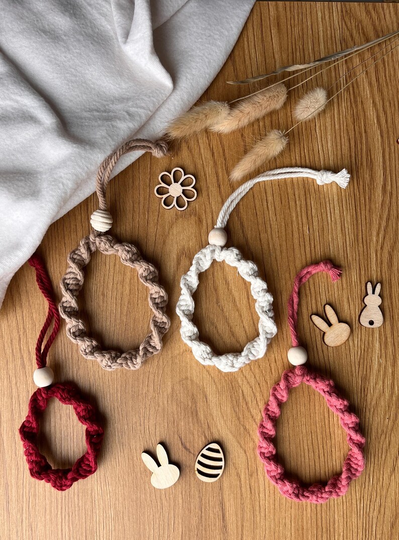 May include: Four macrame Easter egg decorations in red, beige, white, and pink, each with a wooden bead and hanging loop. Wooden bunny and egg cutouts are scattered on a wooden surface. The eggs are for Easter.