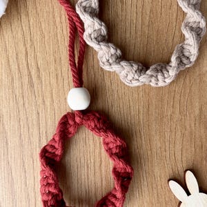 May include: Close-up of two handmade macrame rings. One is a deep red colour, and the other is a neutral beige. The red ring is attached to a red cord with a white wooden bead. The rings are on a wooden surface.