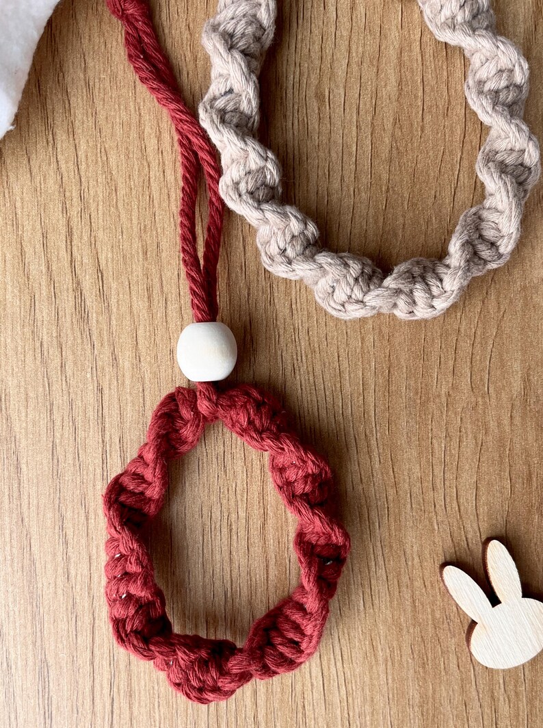 May include: Close-up of two handmade macrame rings. One is a deep red colour, and the other is a neutral beige. The red ring is attached to a red cord with a white wooden bead. The rings are on a wooden surface.