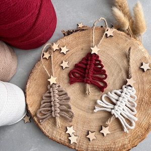 Può includere: Tre addobbi per l'albero di Natale in macramè fatti a mano in rosso, beige e bianco. Gli addobbi sono esposti su una fetta di legno con stelle di legno sparse intorno.