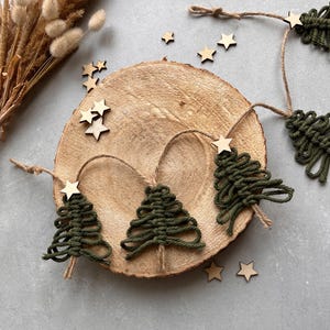 May include: A garland of green yarn Christmas trees with wooden star accents on a wooden slice background.