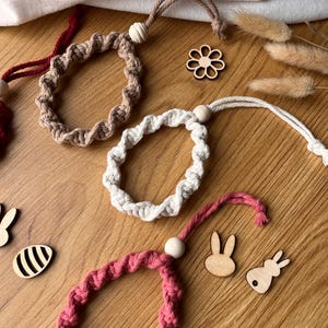 May include: Several handmade macrame ornaments in beige, white, and pink. Each has a looped design, a wooden bead, and a hanging cord. Wooden Easter-themed cutouts, including bunnies and an egg, are also visible, all on a wooden surface.