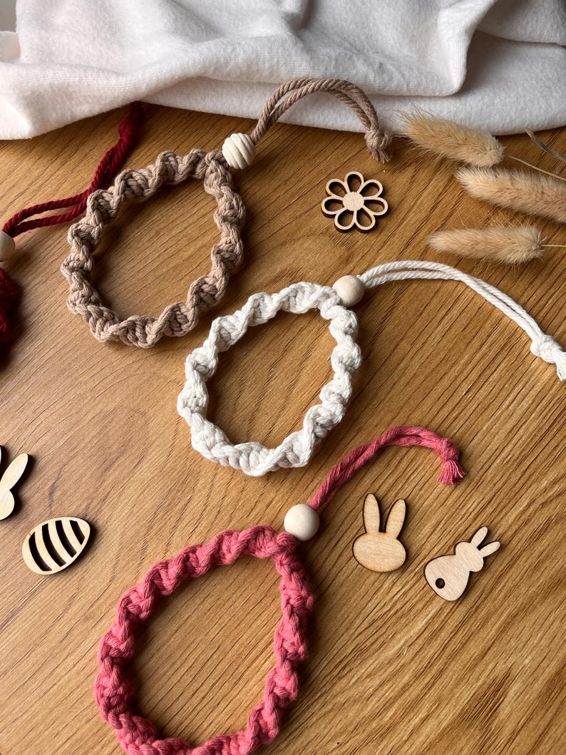 May include: Several handmade macrame ornaments in beige, white, and pink. Each has a looped design, a wooden bead, and a hanging cord. Wooden Easter-themed cutouts, including bunnies and an egg, are also visible, all on a wooden surface.