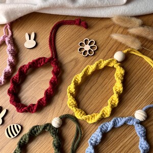 May include: A selection of colourful, handmade macrame bracelets in red, yellow, green, and blue, each with a wooden bead accent. Wooden bunny and flower cutouts are scattered around the bracelets on a wooden surface. The bracelets are a unique accessory.