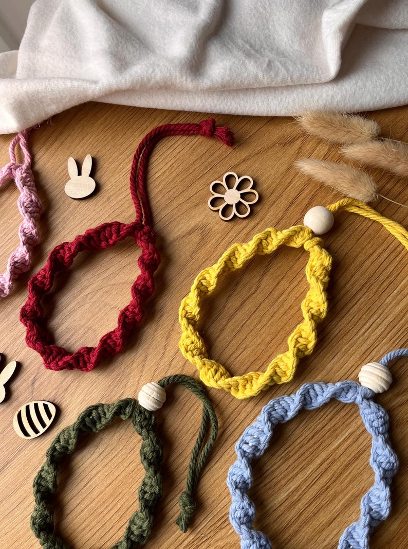 May include: A selection of colourful, handmade macrame bracelets in red, yellow, green, and blue, each with a wooden bead accent. Wooden bunny and flower cutouts are scattered around the bracelets on a wooden surface. The bracelets are a unique accessory.