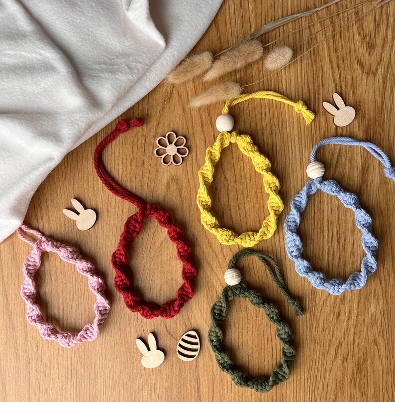 May include: A selection of colourful, handmade Easter egg decorations. The ornaments are in shades of pink, red, yellow, blue, and green, with a wooden bead and a loop for hanging. Wooden bunny and flower cutouts are scattered around.