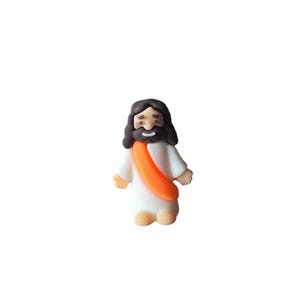 May include: A small figurine of Jesus Christ, depicted with dark brown hair and a beard, wearing a white robe and an orange sash. The figure has a smiling expression and is standing upright.