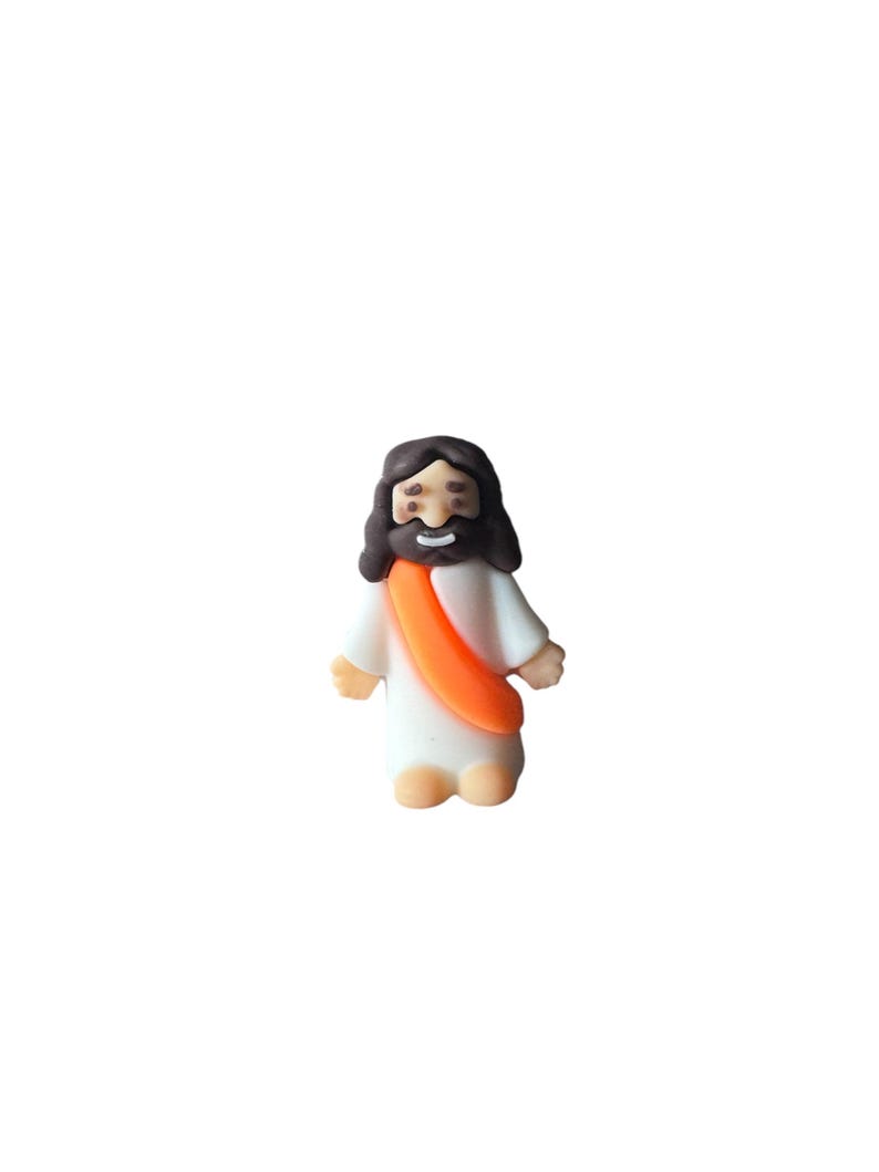 May include: A small figurine of Jesus Christ, depicted with dark brown hair and a beard, wearing a white robe and an orange sash. The figure has a smiling expression and is standing upright.
