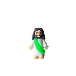 May include: A small, cartoon-style figurine of a religious figure. The figure has dark brown hair and a beard, wearing a white robe with a green sash. The figurine has a smiling expression and is standing.