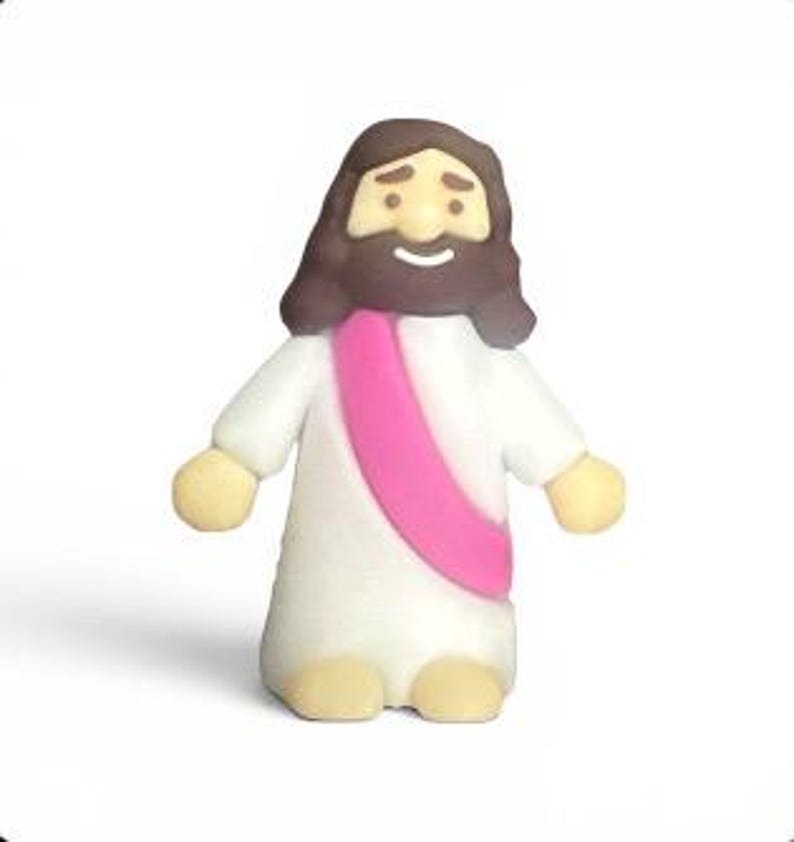 May include: A small, cartoon-like figurine of a religious figure. The figure has a white robe, a pink sash, and brown hair and beard. The figure's arms and feet are a light tan color. The figure is smiling.