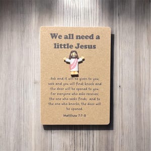 May include: A small, rectangular card with the text "We all need a little Jesus" and a small Jesus figurine. The card also includes a Bible verse from Matthew 7:7-8. The card is a light brown colour.