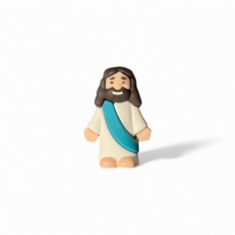 May include: A small figurine of a religious figure, depicted with dark brown hair and a beard. The figure wears a white robe with a teal sash. The figurine has a smiling expression and is standing upright.