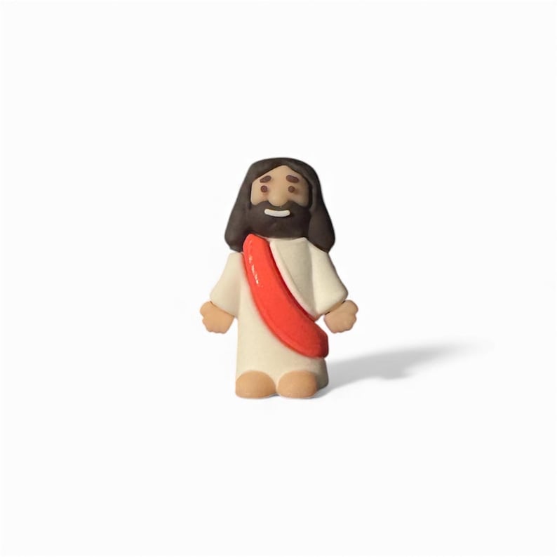 May include: A small figurine of Jesus Christ, depicted with dark brown hair and a beard. He wears a white robe with a red sash. The figurine has a smiling expression and is standing upright.