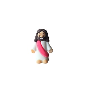 May include: A small figurine of Jesus Christ, depicted with dark brown hair and a beard. He wears a white robe with a pink sash. The figure has a smiling expression and is standing upright.