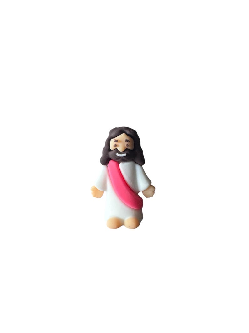 May include: A small figurine of Jesus Christ, depicted with dark brown hair and a beard. He wears a white robe with a pink sash. The figure has a smiling expression and is standing upright.