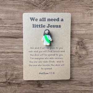 May include: A small, light brown card with the text "We all need a little Jesus" and a small figurine of Jesus. The card also includes a Bible verse from Matthew 7:7-8.
