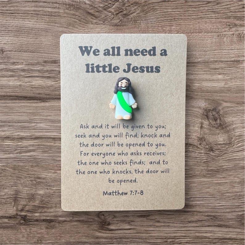 May include: A small, light brown card with the text "We all need a little Jesus" and a small figurine of Jesus. The card also includes a Bible verse from Matthew 7:7-8.