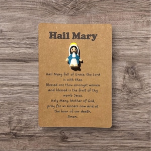 May include: A brown card with the text "Hail Mary" and a prayer. A small figurine of the Virgin Mary is attached. The figurine is wearing a blue and white robe with a yellow halo.