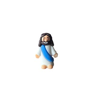 May include: A small figurine of Jesus Christ, depicted with dark brown hair and a beard. He wears a white robe with a blue sash. The figurine has a smiling expression and is standing upright.