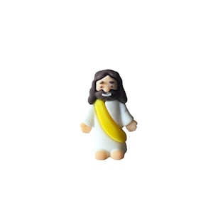 May include: A small, figurine of Jesus Christ, depicted with brown hair and a beard, wearing a white robe and a yellow sash. The figure has a smiling expression and is standing upright.