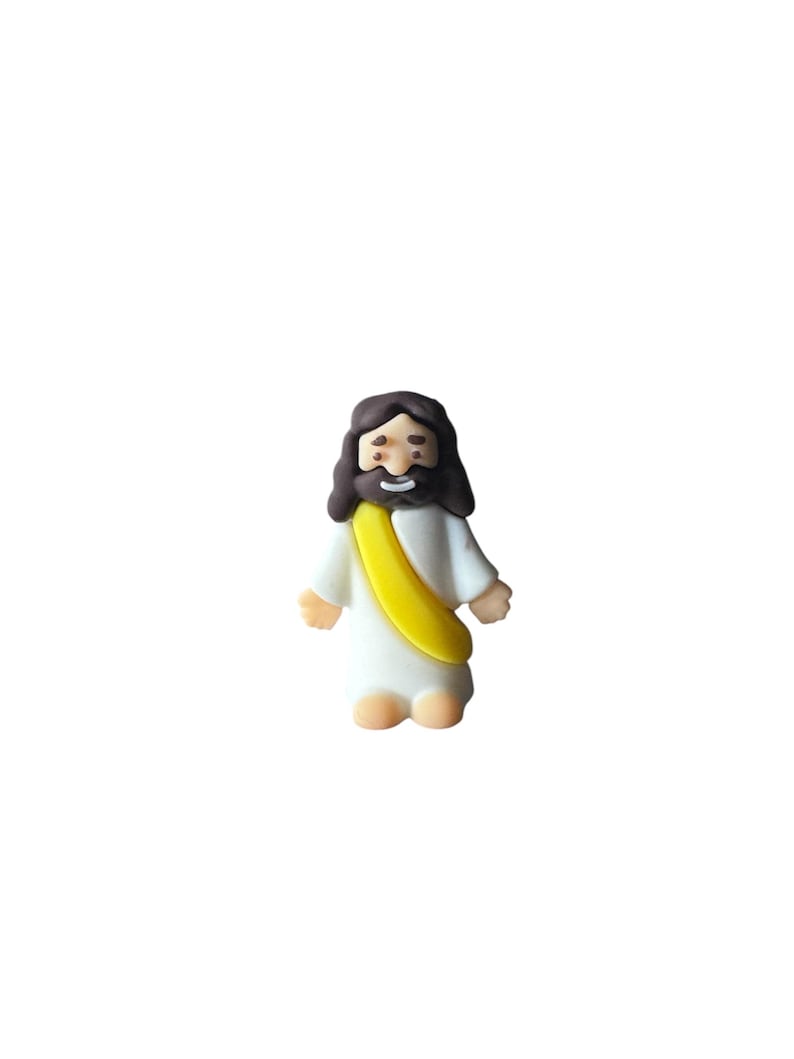 May include: A small, figurine of Jesus Christ, depicted with brown hair and a beard, wearing a white robe and a yellow sash. The figure has a smiling expression and is standing upright.
