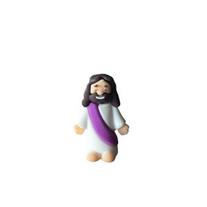 May include: A small, cartoon-style figurine of a religious figure. The figure has dark brown hair and a beard, a white robe, and a purple sash. The figure is smiling and has tan skin.