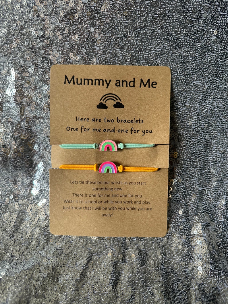 May include: A card with the text "Mummy and Me" and a rainbow design. The card is accompanied by two bracelets with rainbow charms. The text on the card reads "Here are two bracelets, One for me and one for you. Let's tie these on our wrists as you start something new. There is one for me and one for you. Wear it to school or while you work and play. Just know that I will be with you while you are away!"