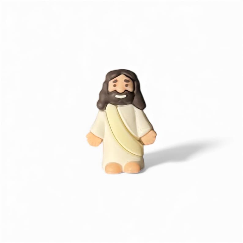 May include: A small, figurine of Jesus Christ, depicted with brown hair and a beard, wearing a white robe with a yellow sash. The figure has a smiling expression and is standing upright.