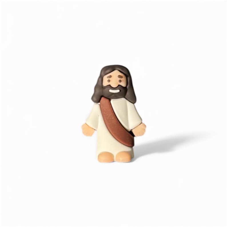 May include: A small, figurine of Jesus Christ, depicted with brown hair and a beard, wearing a white robe and a brown sash. The figure has a smiling expression and is standing on a white surface.